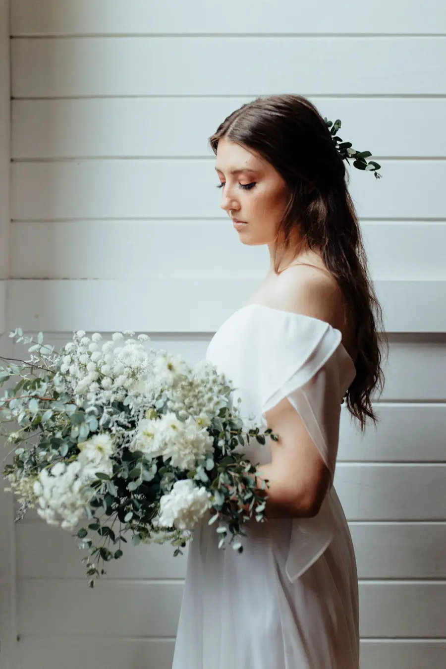 Noiva em vestido minimalista ombro a ombro, segurando buquê de flores brancas e folhagens, em ambiente interno iluminado.