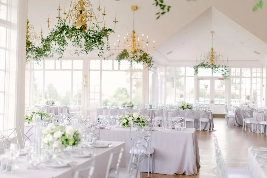 Salão iluminado de casamento com mesas claras, arranjos de flores brancas e lustres dourados com folhagens suspensas.