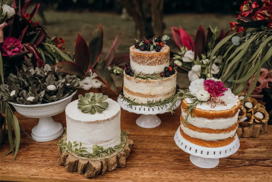Três bolos naked cake decorados com flores e frutas em mesa de casamento simples ao ar livre.