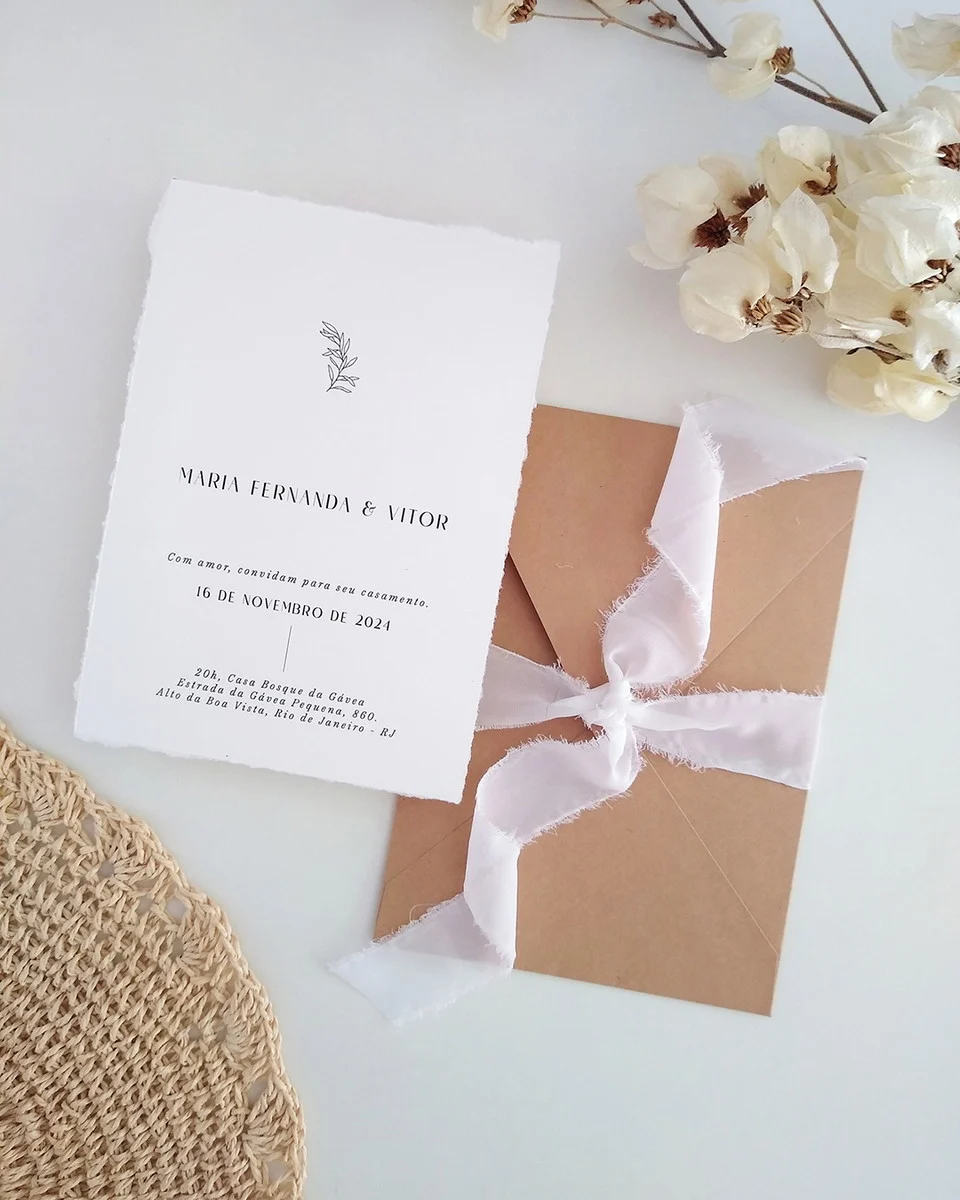 Convite de casamento simples ao ar livre em papel branco com envelope kraft e fita branca, ao lado de flores secas.