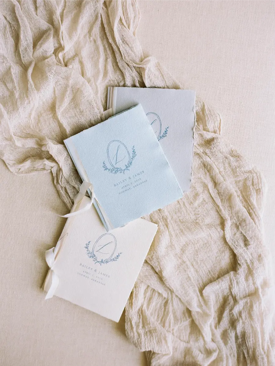 Livretos de votos de casamento em tons de azul e bege com la&ccedil;o de fita, dispostos sobre tecido r&uacute;stico bege.