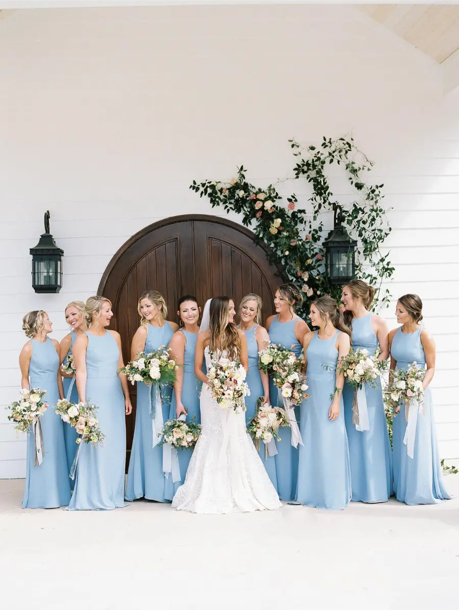 Noiva com madrinhas em vestidos azul serenity posando diante de capela branca.