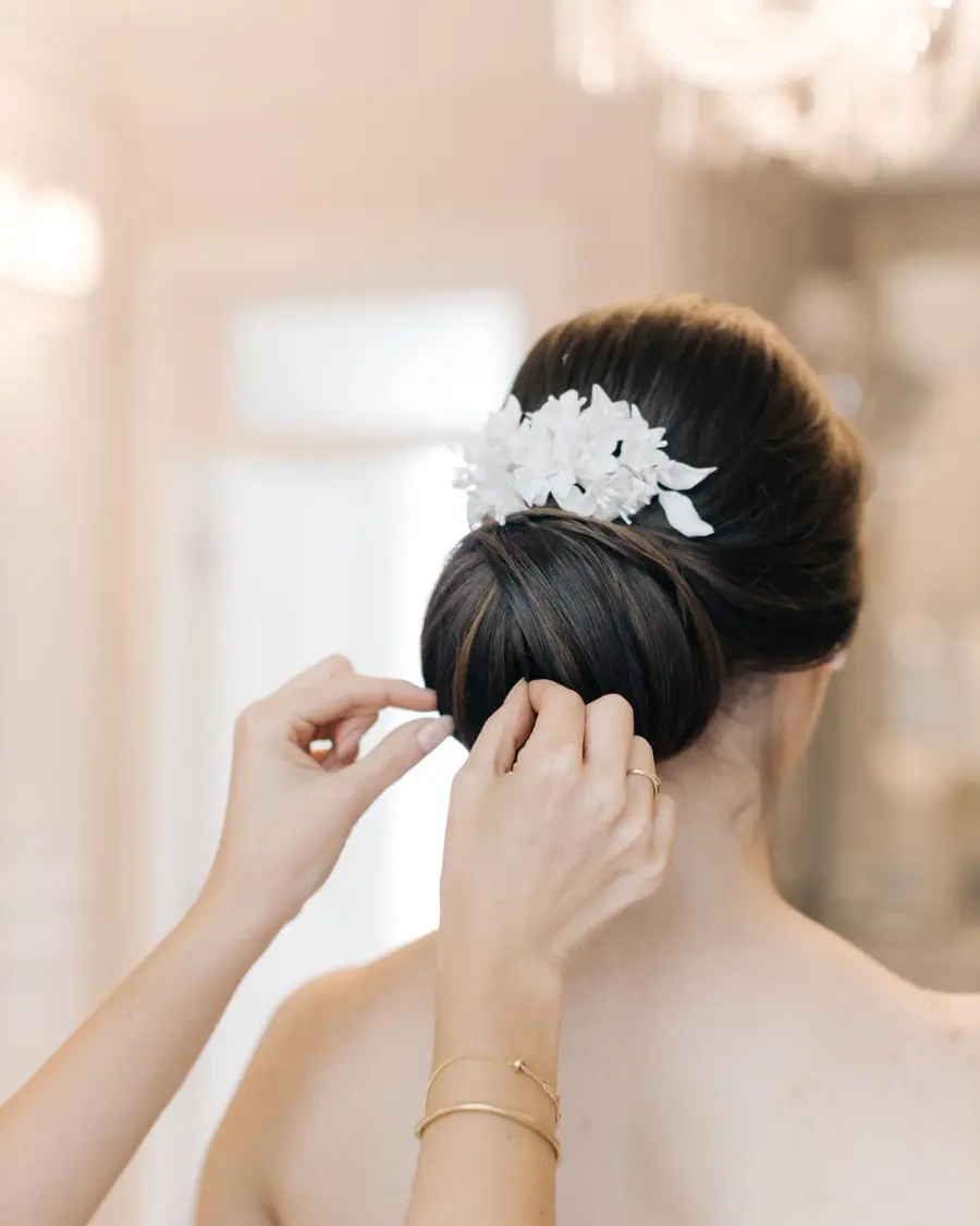 Noiva com coque baixo decorado com flores brancas sendo finalizado pelas mãos da cabeleireira