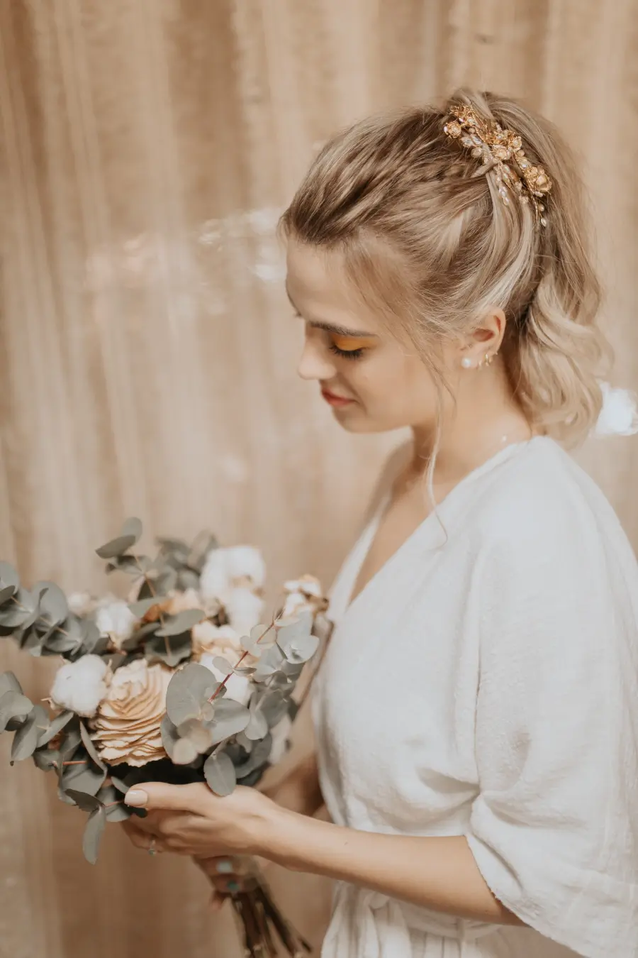 Noiva loira com rabo de cavalo estilizado e acessório dourado no cabelo, segurando buquê rústico com eucalipto e flores secas.