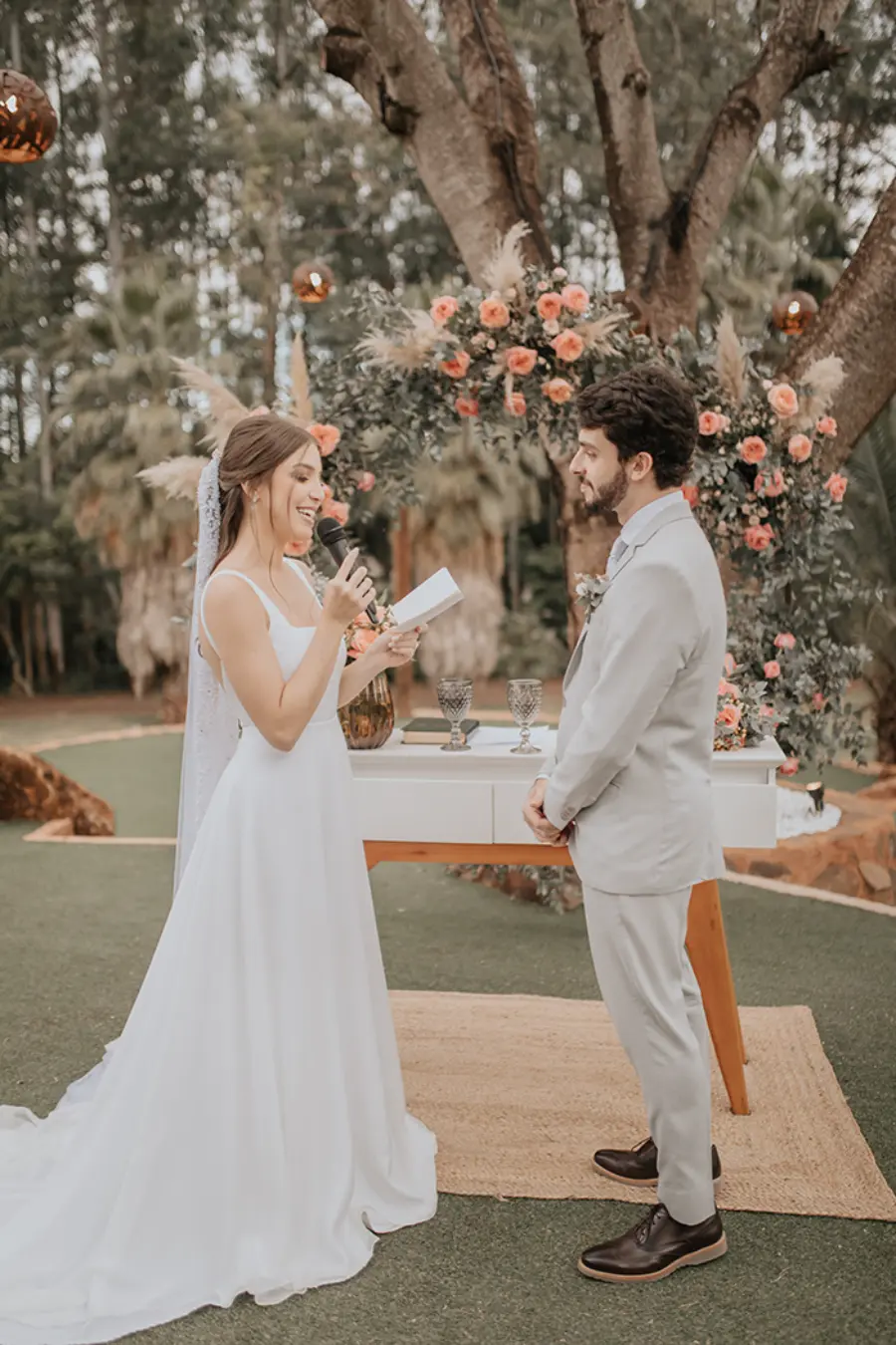 Cena da cerimônia em espaço externo, onde a noiva, de vestido branco, lê seus votos ao noivo diante de um altar decorado com flores.