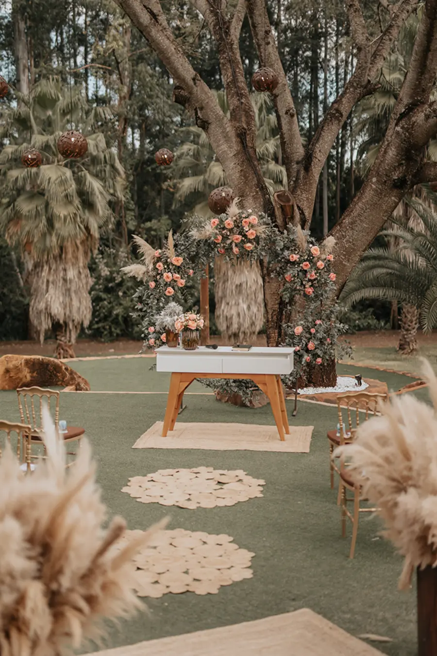 Altar com mesa de madeira clara e arco de flores e folhagens montado no tronco de uma grande árvore