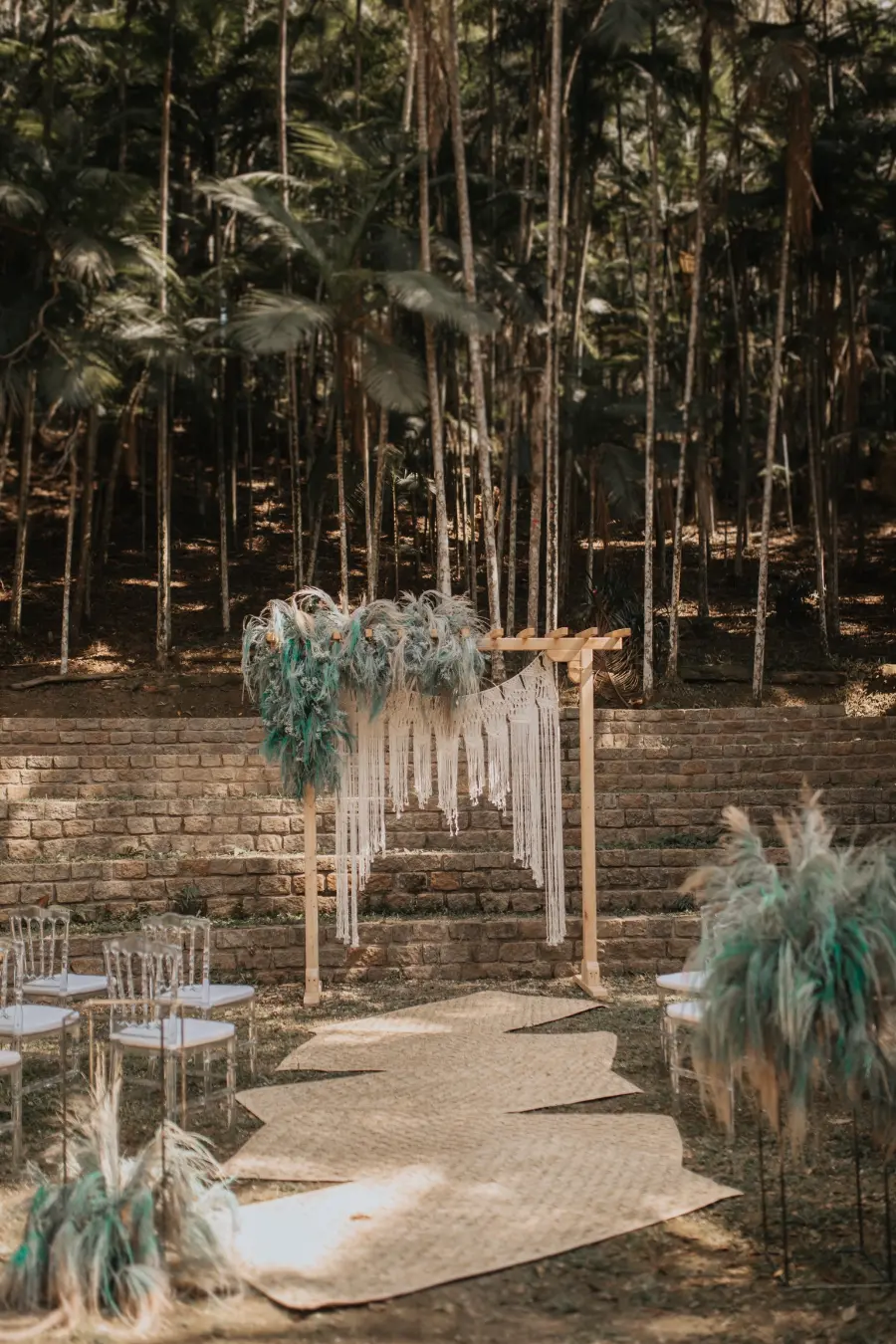 Altar de casamento rústico com arranjos de capim dos pampas e macramê, em meio a uma área arborizada com bancos para os convidados.