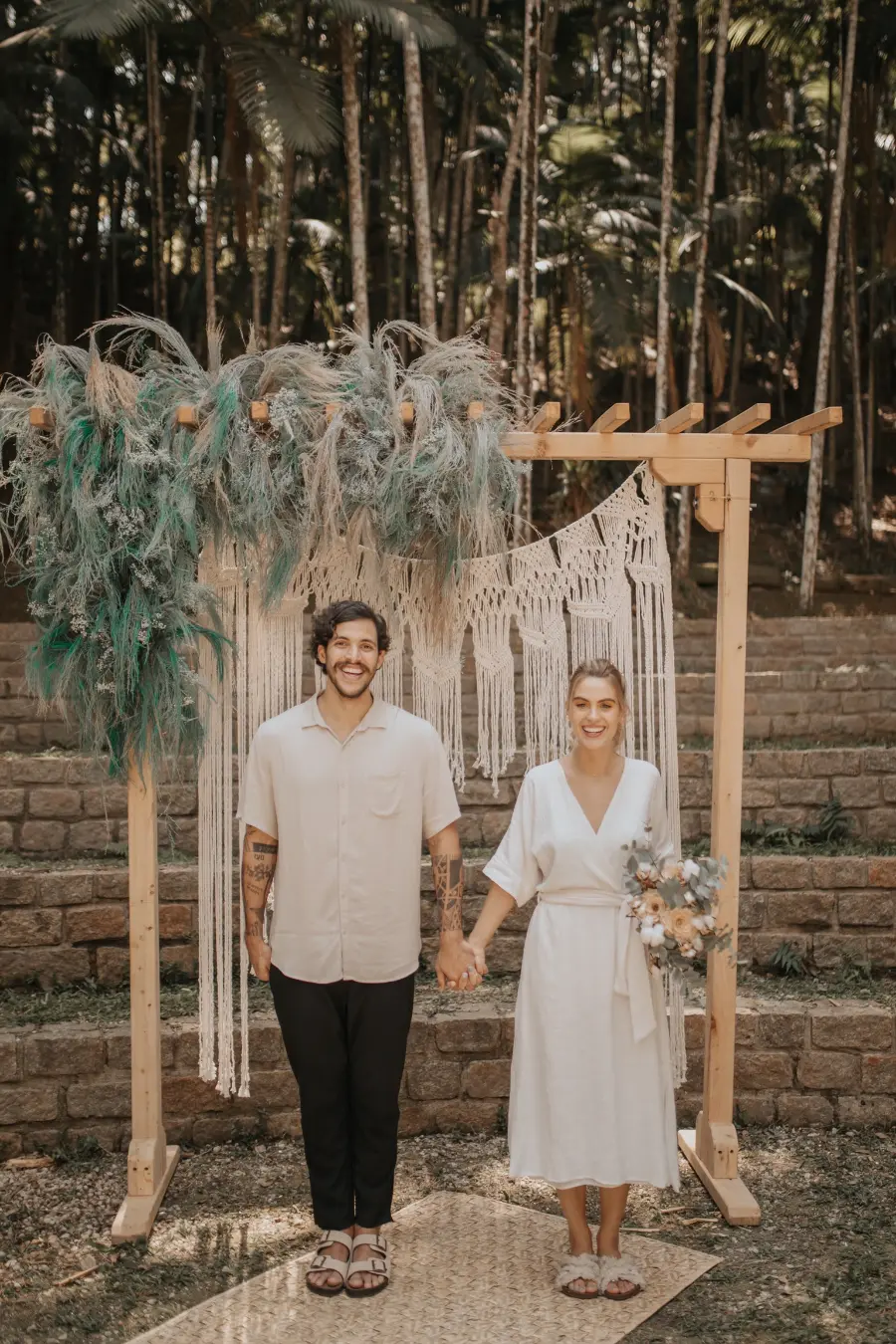 Noivos de mãos dadas em frente a um altar de madeira decorado com macramê e folhagens secas, em ambiente rústico ao ar livre.