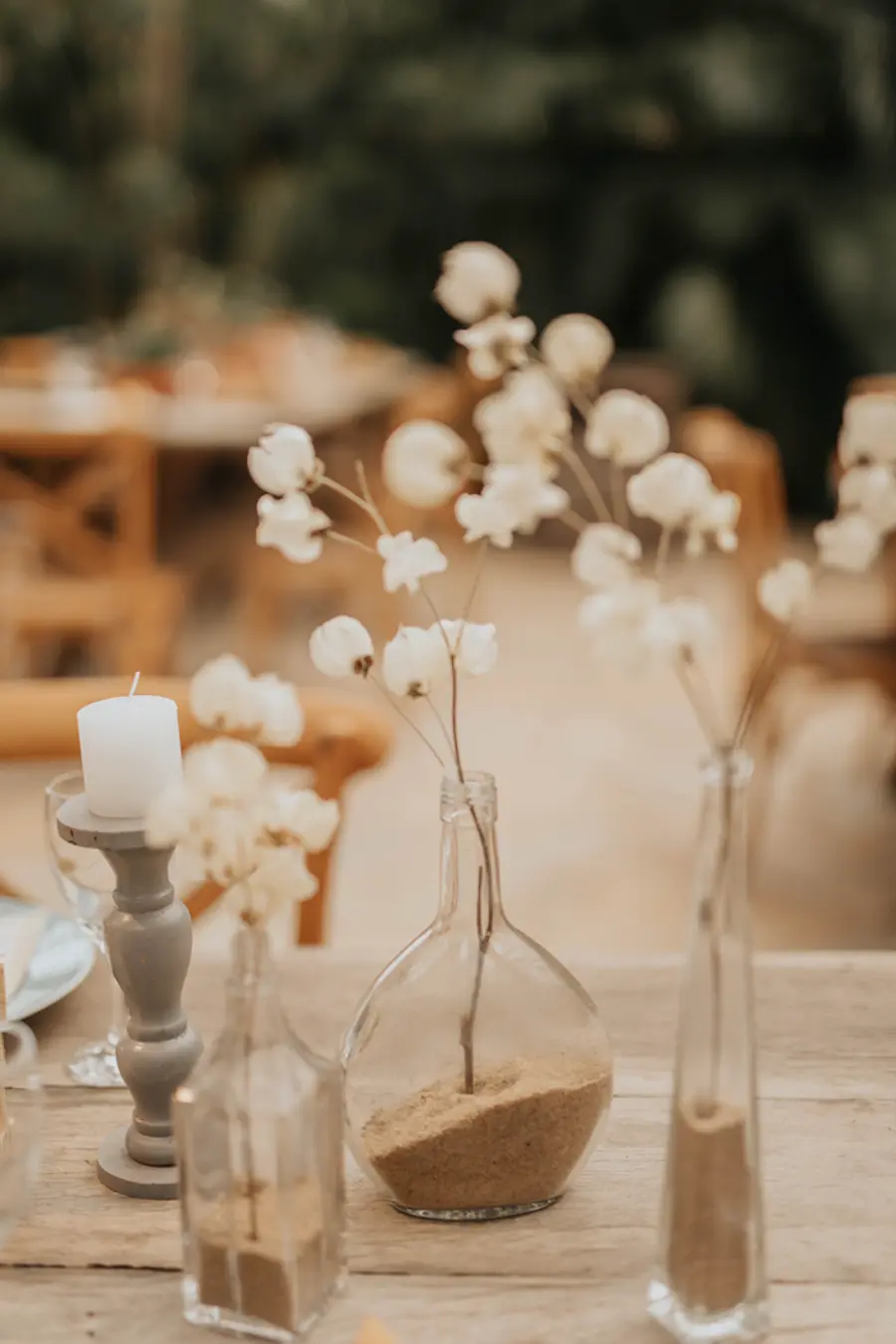 Detalhe de decoração de mesa com garrafas de vidro preenchidas com areia e ramos delicados, acompanhados de vela branca.