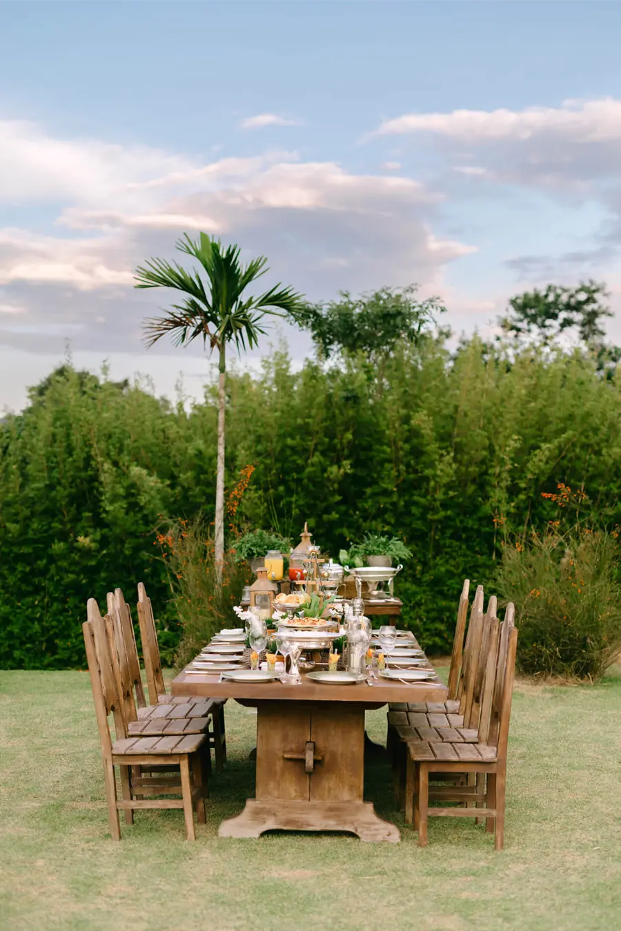 Mesa de madeira comprida com cadeiras no mesmo estilo, decorada com pratos, talheres e alimentos para recepção