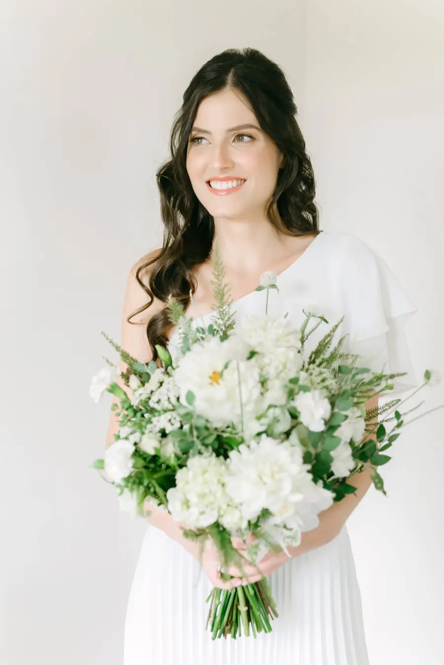Noiva de vestido branco com detalhe em um ombro, segurando buquê volumoso de flores brancas e folhagens, em fundo claro.