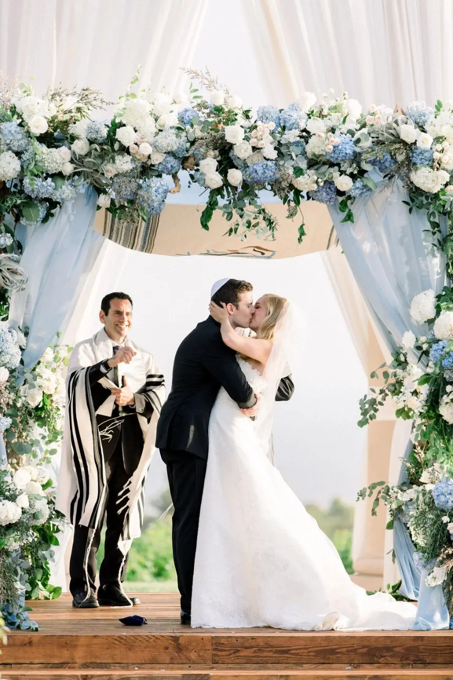 Noivos se beijando em cerim&ocirc;nia sob arco floral com flores azuis e brancas e cortinas em azul serenity.