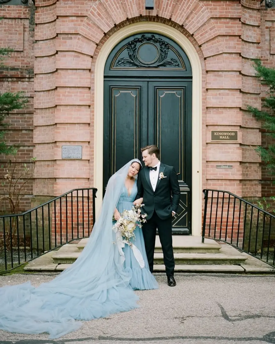 Noiva com vestido azul serenity e v&eacute;u longo ao lado do noivo de terno preto em frente a igreja.