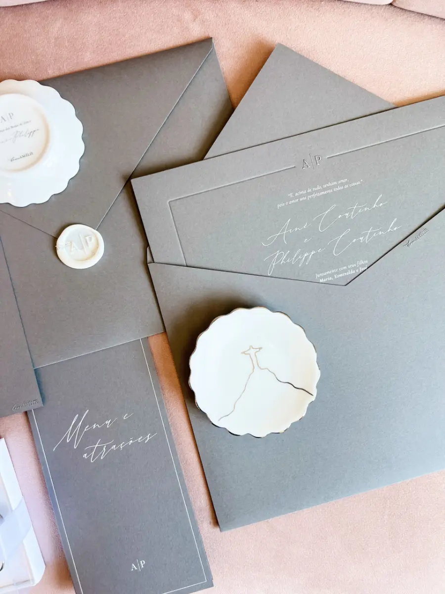 Convite de casamento minimalista em papel cinza com monograma em relevo, acompanhado de prato de porcelana com desenho do Cristo Redentor.
