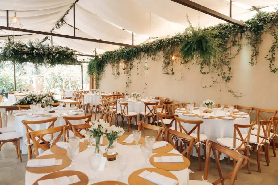 Sal&atilde;o de festa para casamento com teto de lona, cadeiras de madeira cruzada e decora&ccedil;&atilde;o verde suspensa