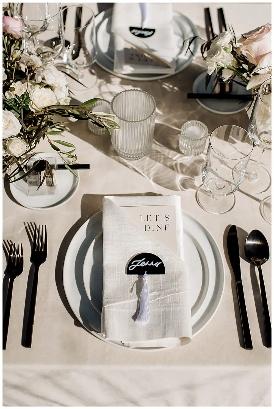 Mesa posta para casamento com arranjos de flores brancas, guardanapo com plaquinha personalizada e talheres pretos.
