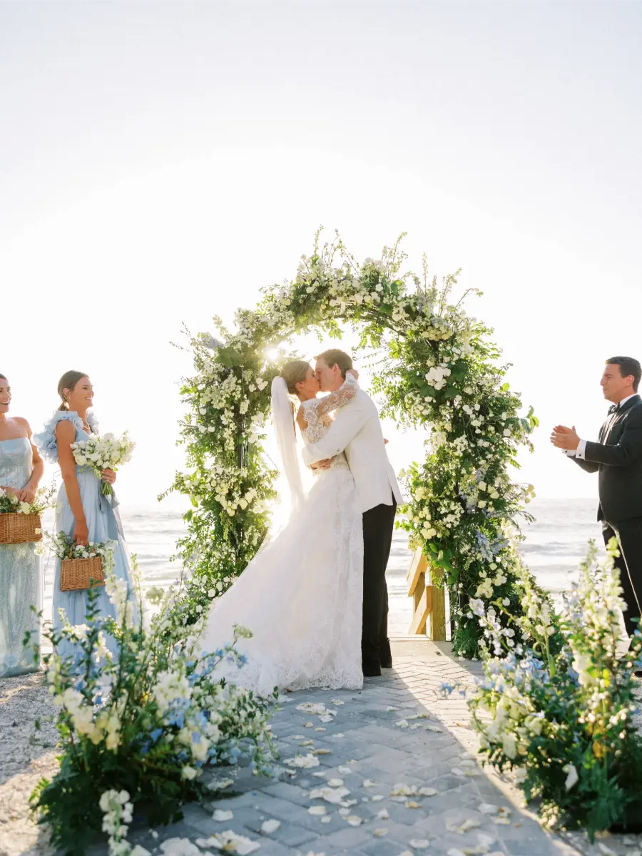 Casal se beijando sob arco de flores brancas e azuis em cerim&ocirc;nia de casamento na praia com decora&ccedil;&atilde;o azul serenity.