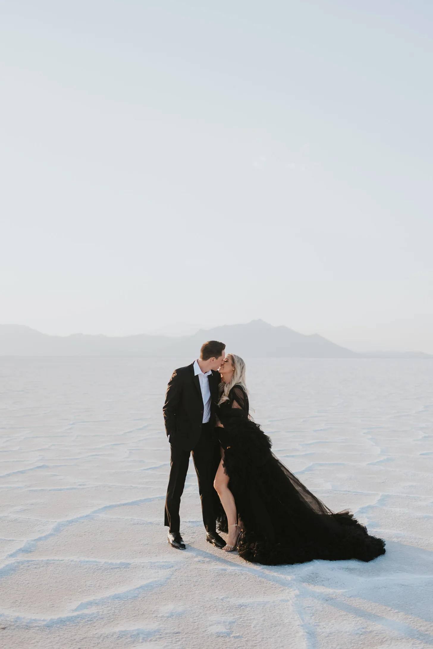 Casal de noivos se beijando em paisagem branca com noiva usando vestido preto volumoso.