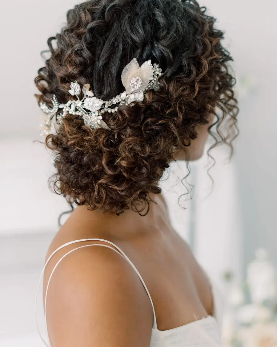 Noiva com cabelo cacheado curto preso em coque baixo, adornado com tiara prateada de flores e pedrarias.