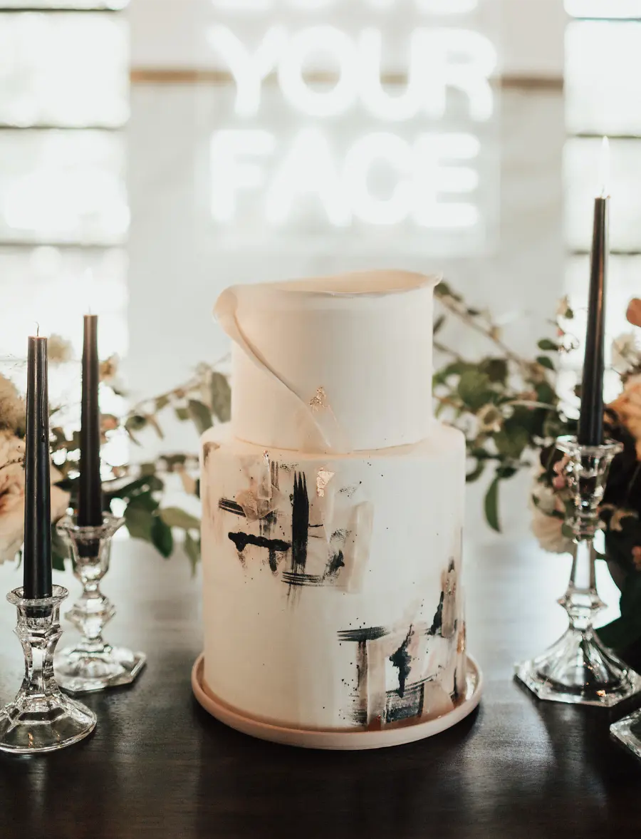 Bolo de casamento art&iacute;stico em branco com pinceladas pretas e douradas, rodeado de velas pretas.