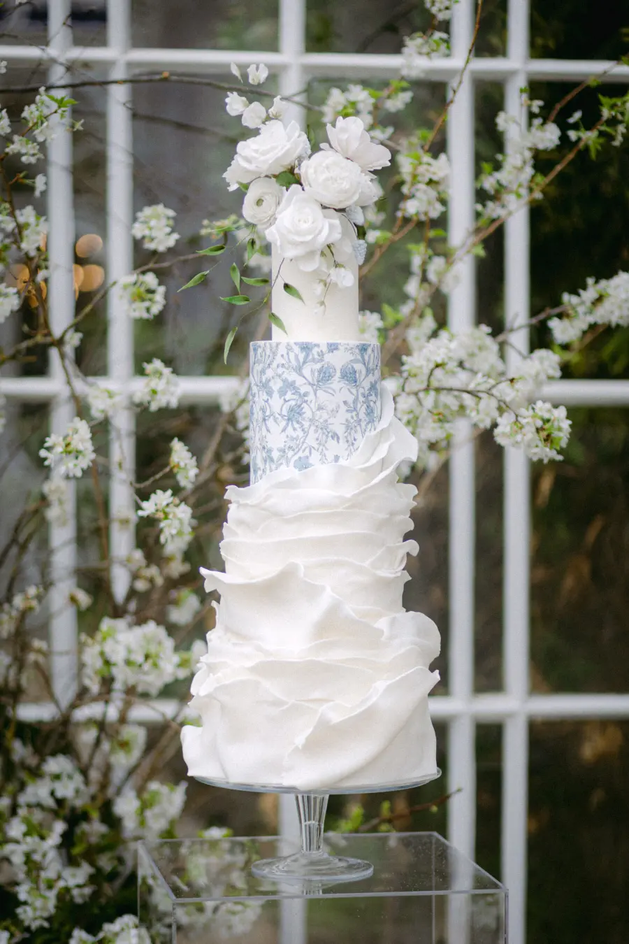 Bolo de casamento alto decorado com flores brancas e camada estampada em azul, em ambiente com flores ao fundo.
