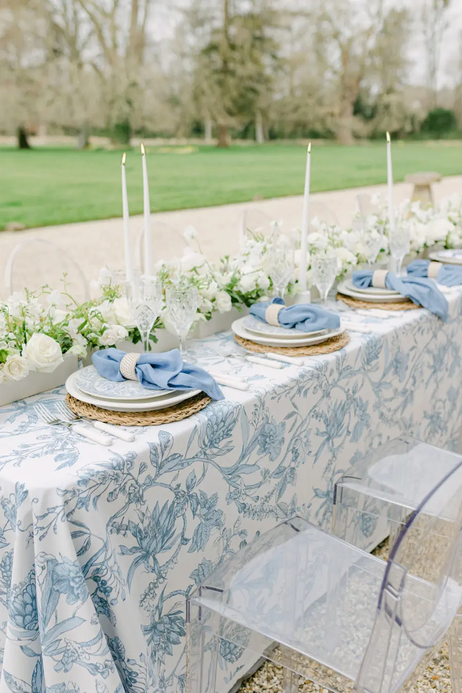 Mesa de casamento ao ar livre com toalha floral azul serenity, guardanapos azuis e arranjo de flores brancas.
