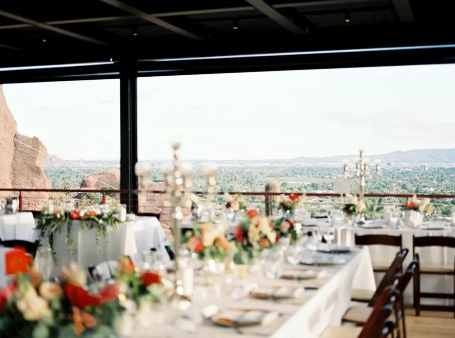 Mesas de casamento decoradas com flores coloridas e candelabros em salão aberto com vista panorâmica para montanhas e vegetação ao fundo.