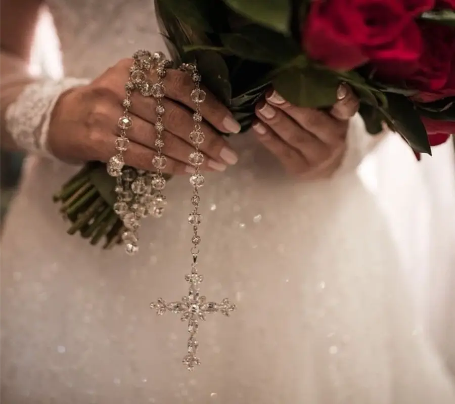 Noiva segurando buquê de rosas vermelhas com terço de cristal prateado.