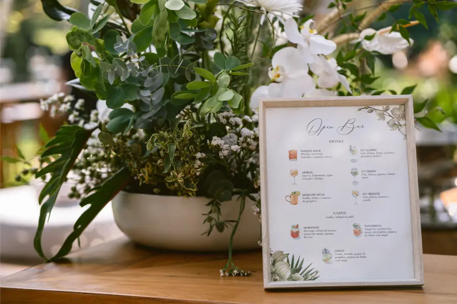Placa em moldura de madeira com cardápio de drinks para open bar, decorando mesa com arranjo floral verde e branco.