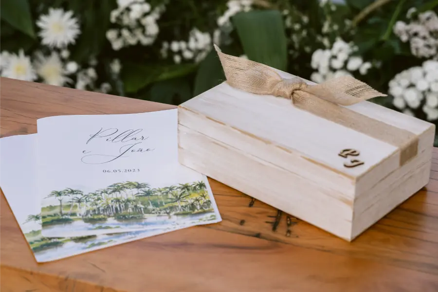 Caixa rústica de madeira com fita de juta ao lado de convite de casamento floral, apoiados sobre mesa de madeira.