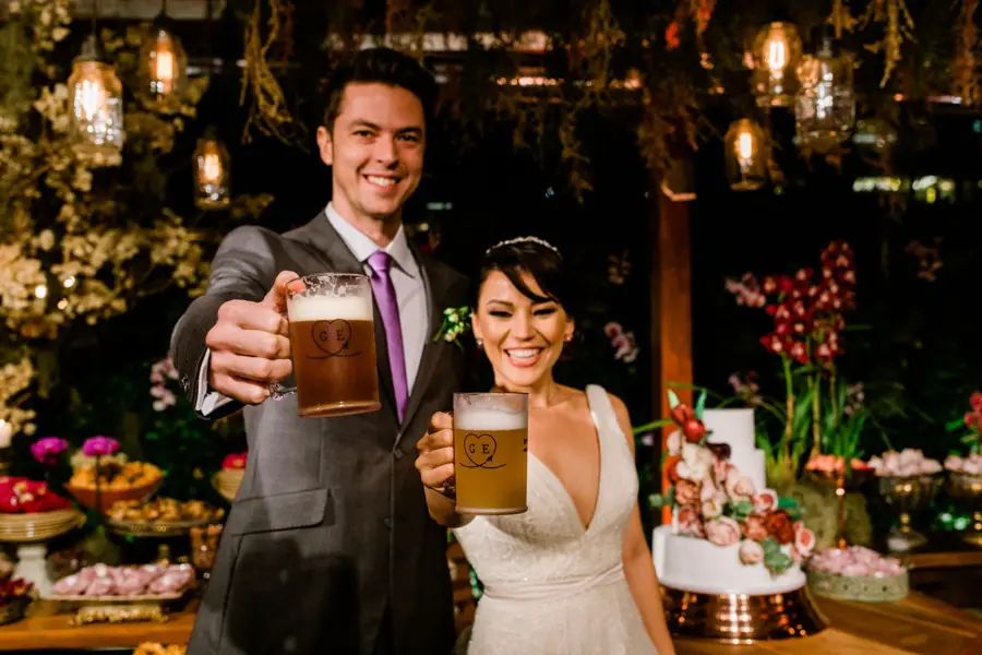 Casal de noivos brindando com canecas de chopp em festa de casamento com decoração rústica e iluminação ambiente