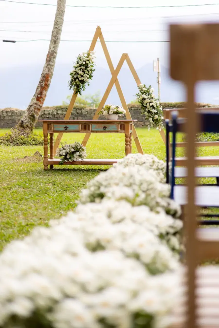 Altar de casamento simples ao ar livre com estrutura triangular de madeira decorada com flores brancas.