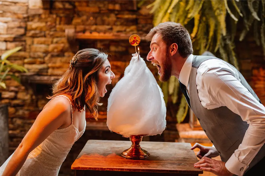 Noiva e noivo se divertem encarando um algodão-doce gigante apoiado em suporte dourado durante a recepção de casamento.