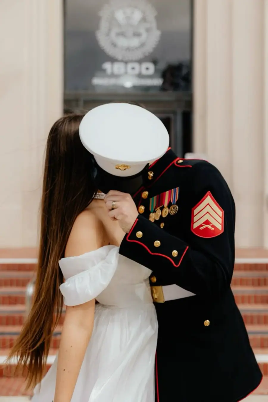 Militar com uniforme preto e vermelho da Marinha dos EUA beija a noiva em frente a uma escadaria.
