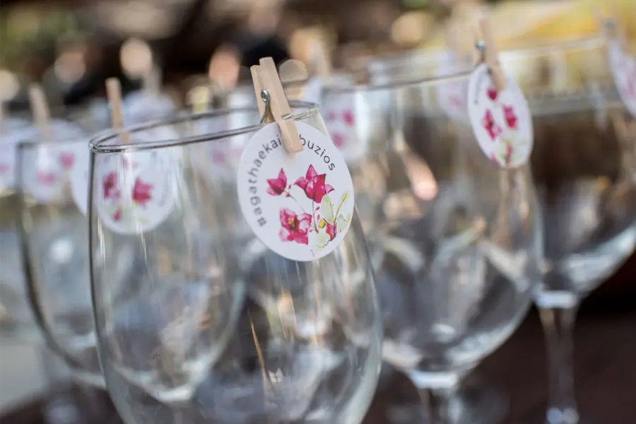 Taças de vinho decoradas com tags redondas de flores cor-de-rosa, presas com mini pregadores de madeira, usadas como identificação em festa de casamento.