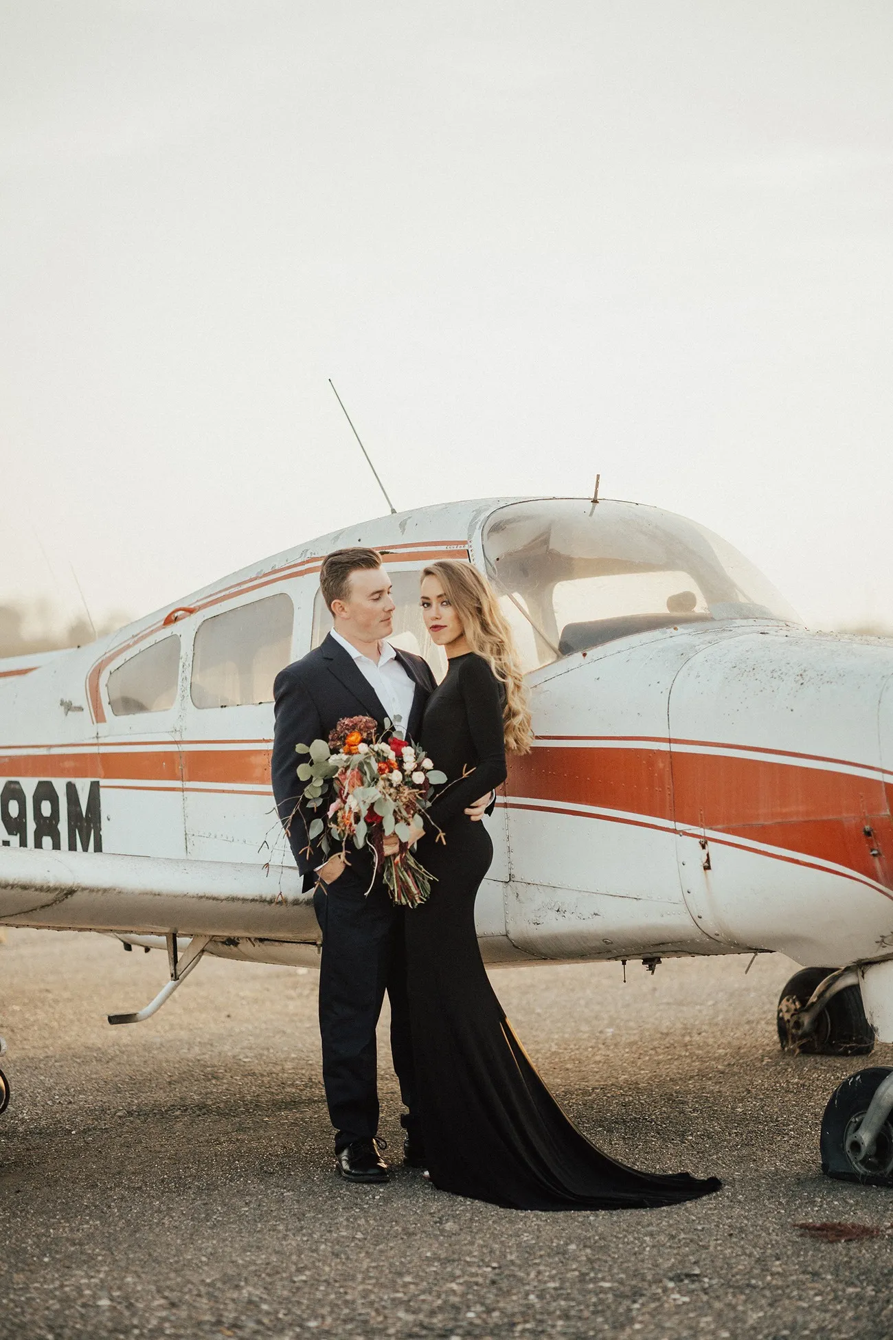 Casal de noivos posando em frente a avi&atilde;o, com noiva em vestido preto e buqu&ecirc; colorido.