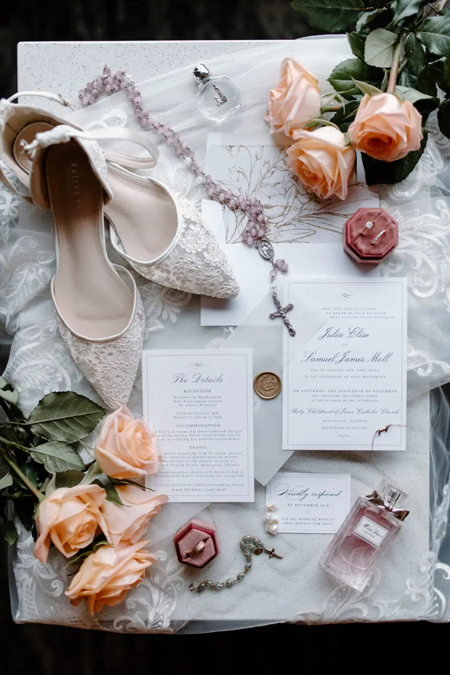 Flat lay com terço rosa, convites de casamento, sapatos de noiva, flores e anéis.