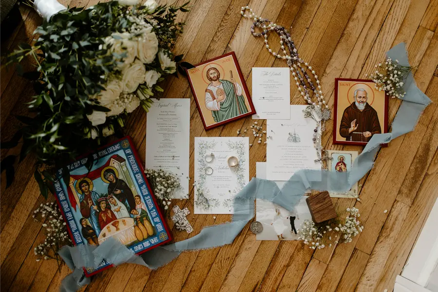 Terço com imagens de santos, convites e flores sobre superfície de madeira.