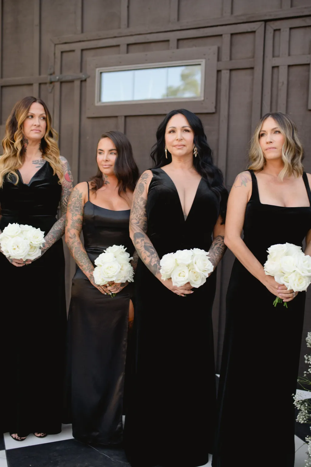 Madrinhas de casamento com vestidos pretos longos segurando buqu&ecirc;s de rosas brancas, em frente a parede de madeira.
