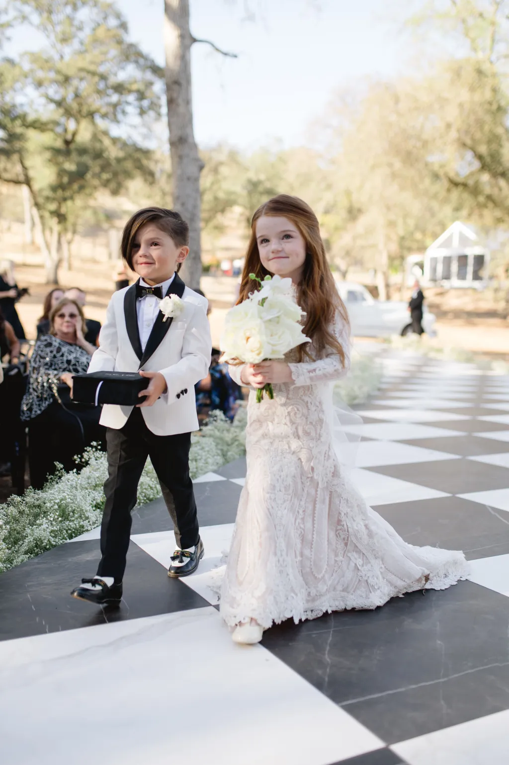 Crian&ccedil;as entrando no casamento: pajem com terno branco e gravata borboleta preta e daminha com vestido rendado claro, segurando buqu&ecirc; de flores brancas.