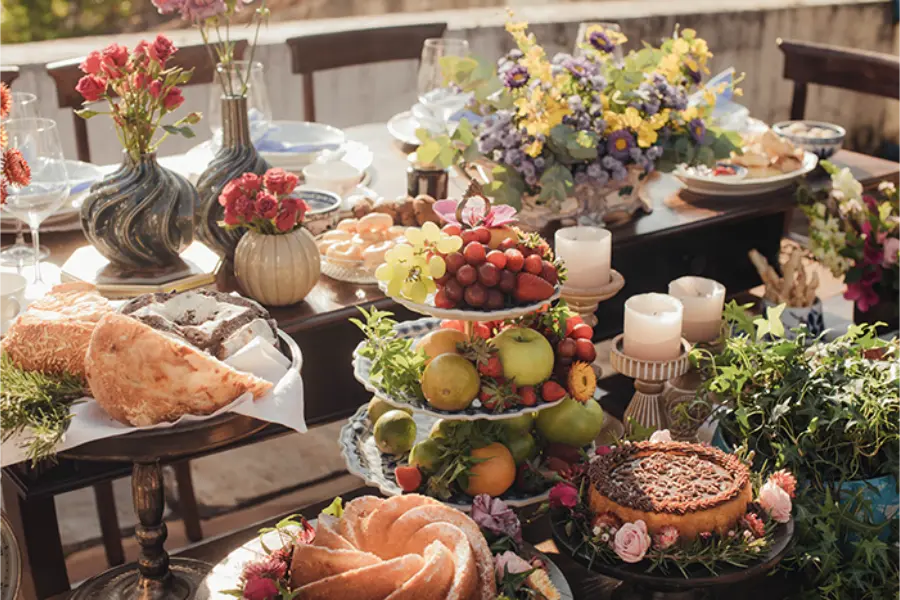 Mesa de casamento simples ao ar livre decorada com flores, frutas e bolos dispostos em diferentes alturas.