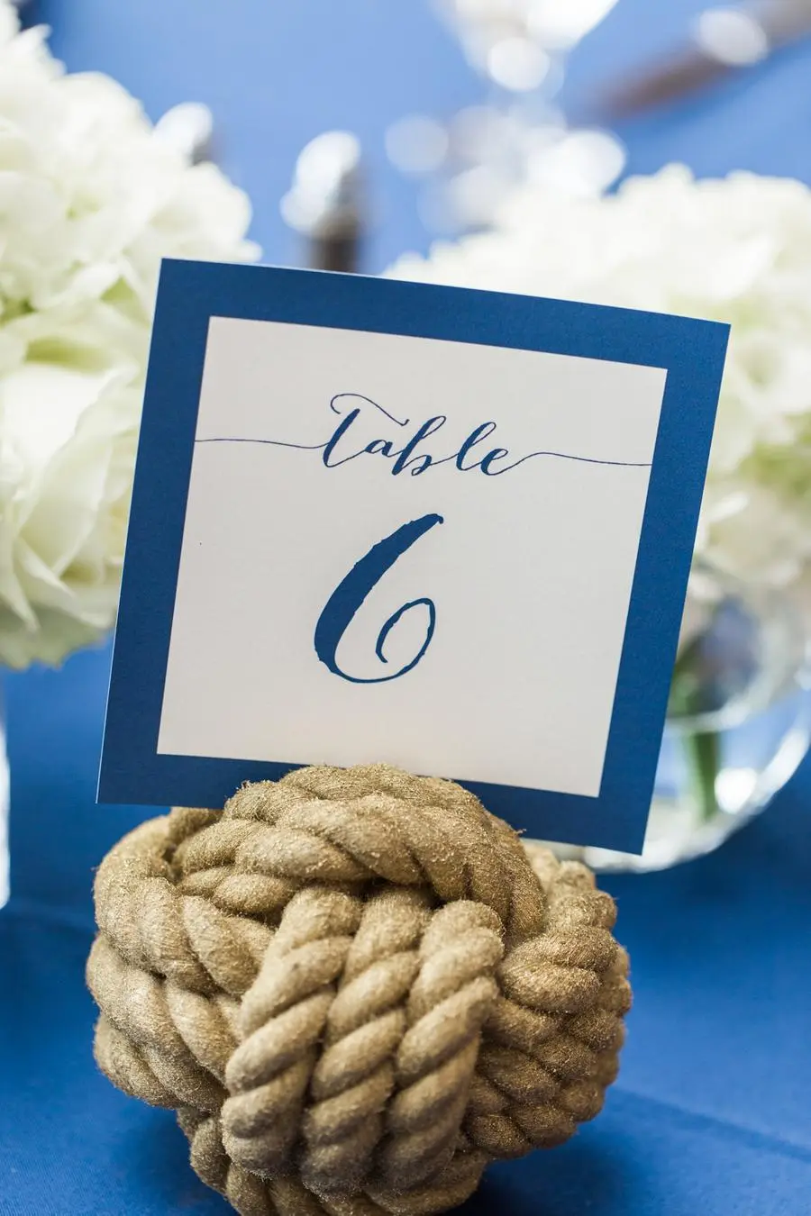 Marcador de mesa azul com n&uacute;mero seis apoiado em n&oacute; n&aacute;utico dourado sobre toalha azul.