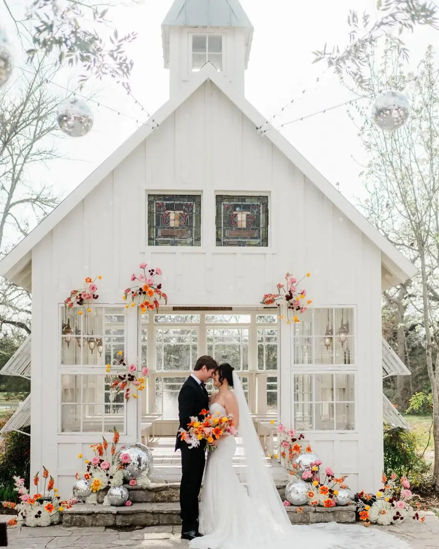 Casal de noivos em frente a capela branca decorada com flores coloridas e detalhes modernos