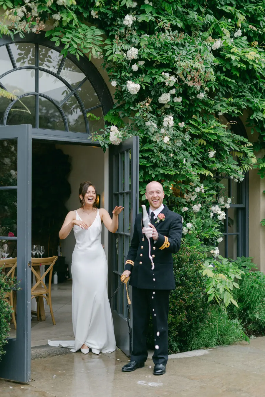 Noivo em uniforme militar abre garrafa de espumante com espada ao lado da noiva sorridente.
