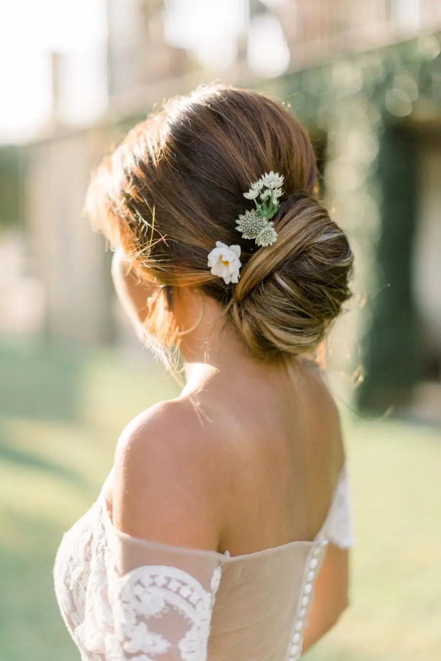 Noiva de costas com coque baixo adornado por flores brancas e verdes em ambiente externo.