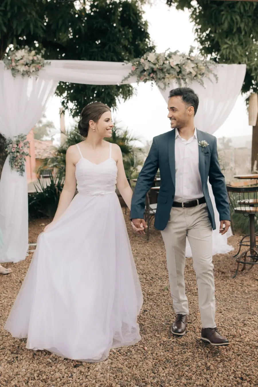 Casal de noivos sorridentes caminhando de mãos dadas em cerimônia ao ar livre, sob arco decorado com flores brancas e rosas.