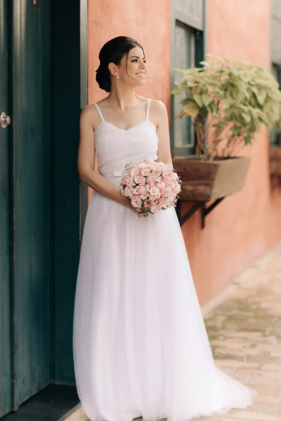 Noiva sorridente em vestido branco simples de alças finas, com buquê de rosas cor-de-rosa, encostada em parede rústica.