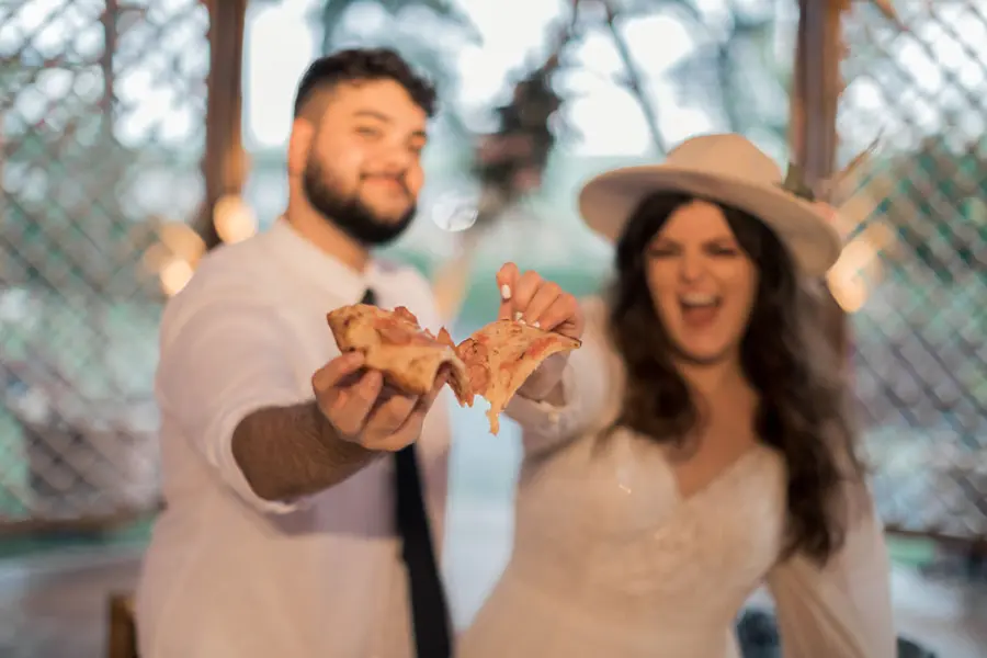 Casal de noivos segurando fatias de pizza como parte do cardápio descontraído.