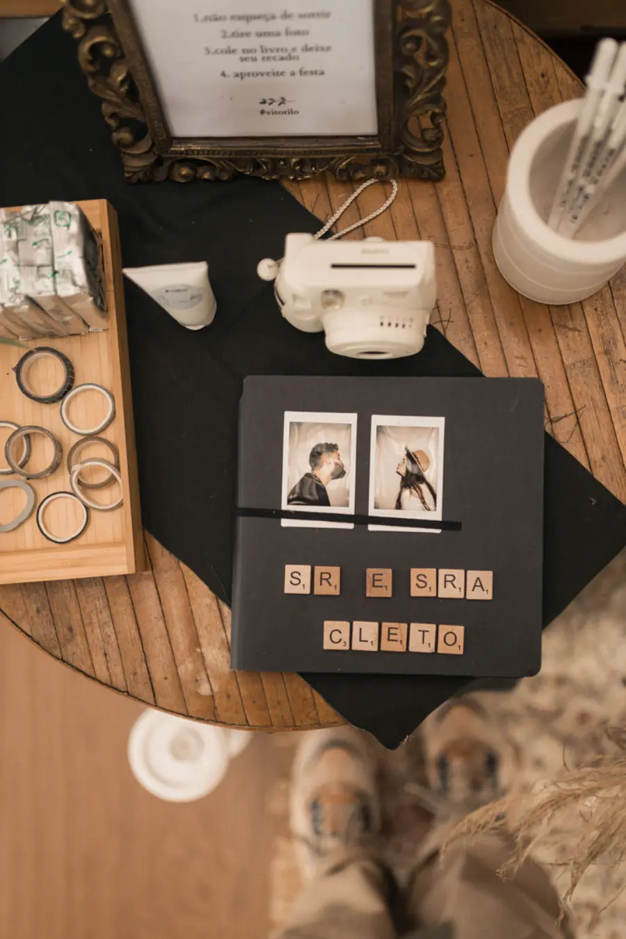 Mesa com câmera instantânea e álbum preto decorado com letras que formam “Sr. e Sra. Cleto”, acompanhado de fotos coladas na capa.