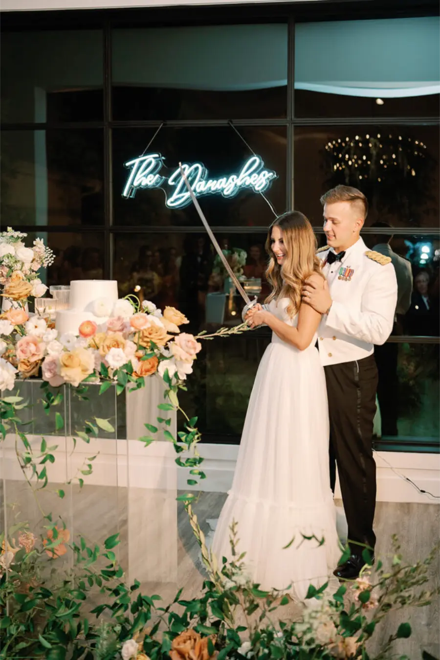 Casal corta o bolo de casamento com espada militar, rodeado por flores e decora&ccedil;&atilde;o elegante.